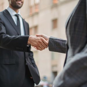 Two business professionals in suits shaking hands on a city street, close-up view of the handshake.